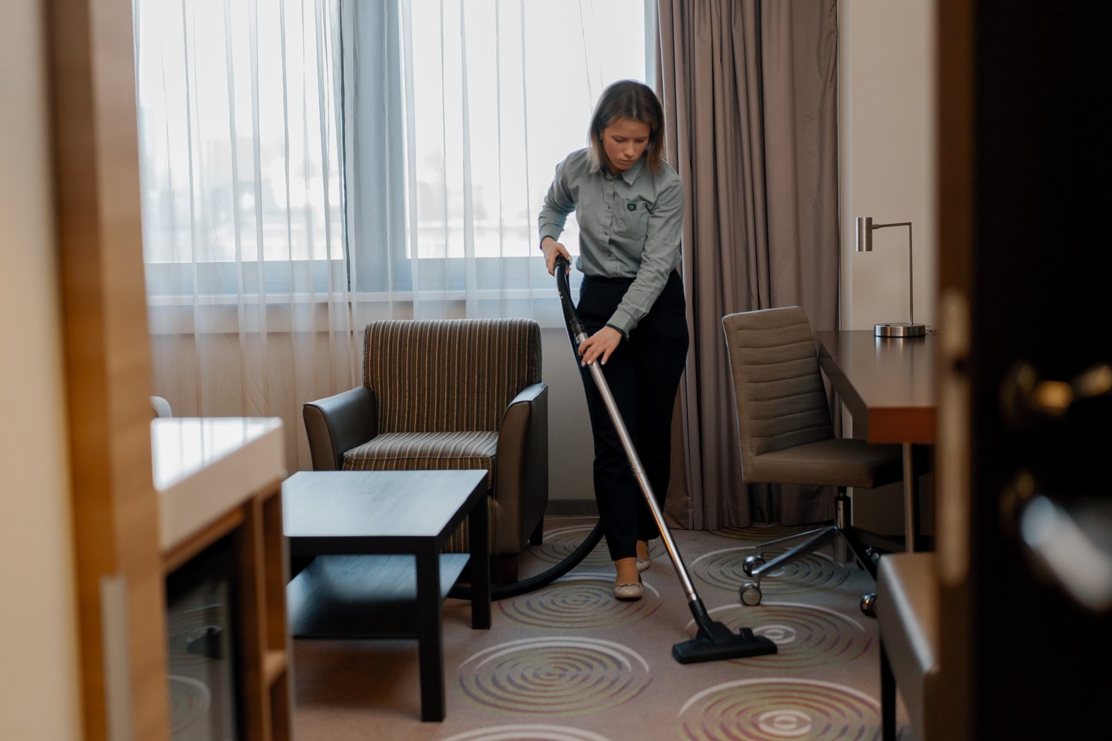 A diligent maid in uniform carefully vacuums the carpet while cleaning hotel room